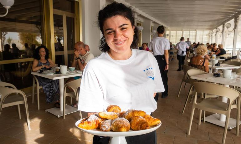 Cameriera sorridente con vassoio di cornetti in un caffè affollato.
